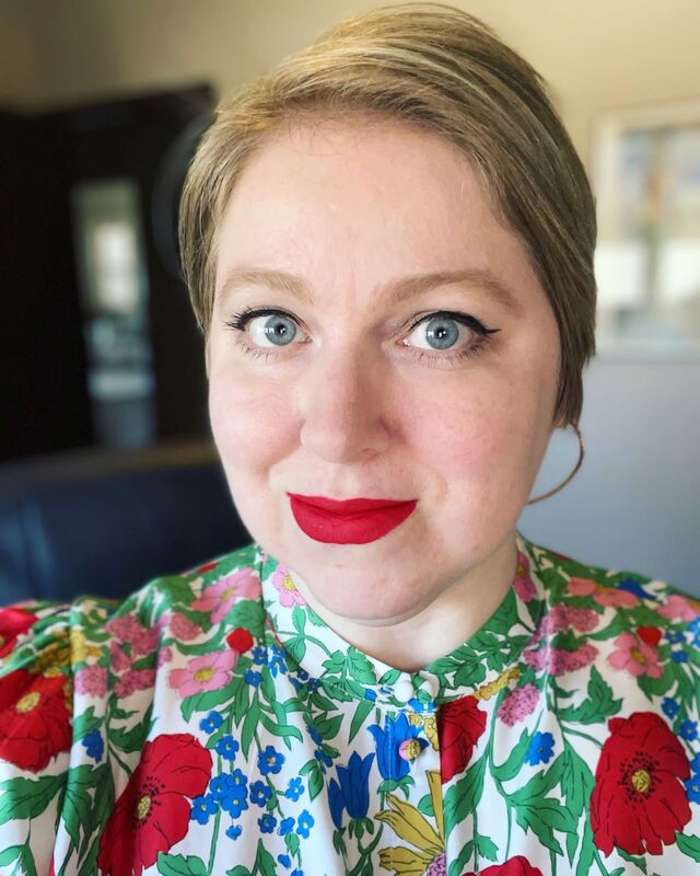 Selfie of Rachel, a white woman in a floral top with short blonde hair, blue eyes, and bright red lipstick