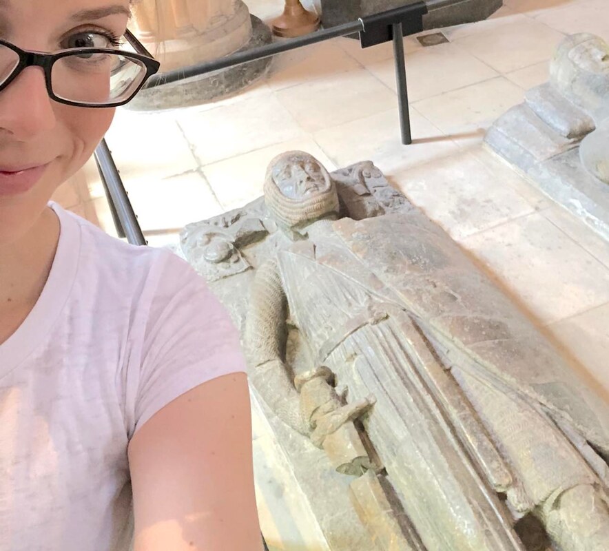 A selfie of Kristin (A blonde woman with black glasses and a light t-shirt) with the effigy of William Marshal, a stone figure of a knight with a shield and sword
