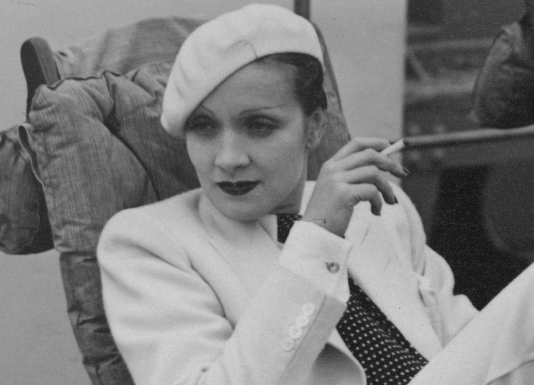 Black and white photograph of Dietrich wearing a white suit and beret and holding a cigarette