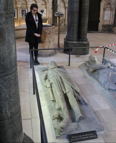 Christine (a woman with dark hair in a braid, wearing a dark blue coat), looking down at the stone knight effigy of William Marshal