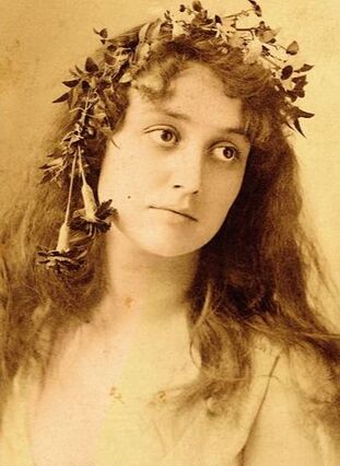 Sepia colored image of a young woman with long dark wavy hair. she has a flower crown on, large dark eyes, and is looking off to her left