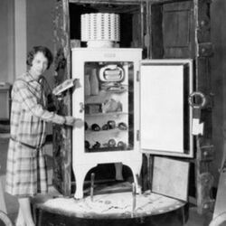 Black and white photograph of a woman in a plaid dress with short hair showing off and old white refrigerator