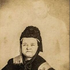 Sepia photograph of Mary Todd Lincoln as an older woman appearing to have a ghost who looks like Abraham Lincoln standing behind her